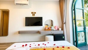 Habitación de hotel con cama, televisión de pantalla plana, mesita pequeña con una cesta y una taza rosa, aire acondicionado instalado en la pared, y ventana grande con vistas a palmeras.