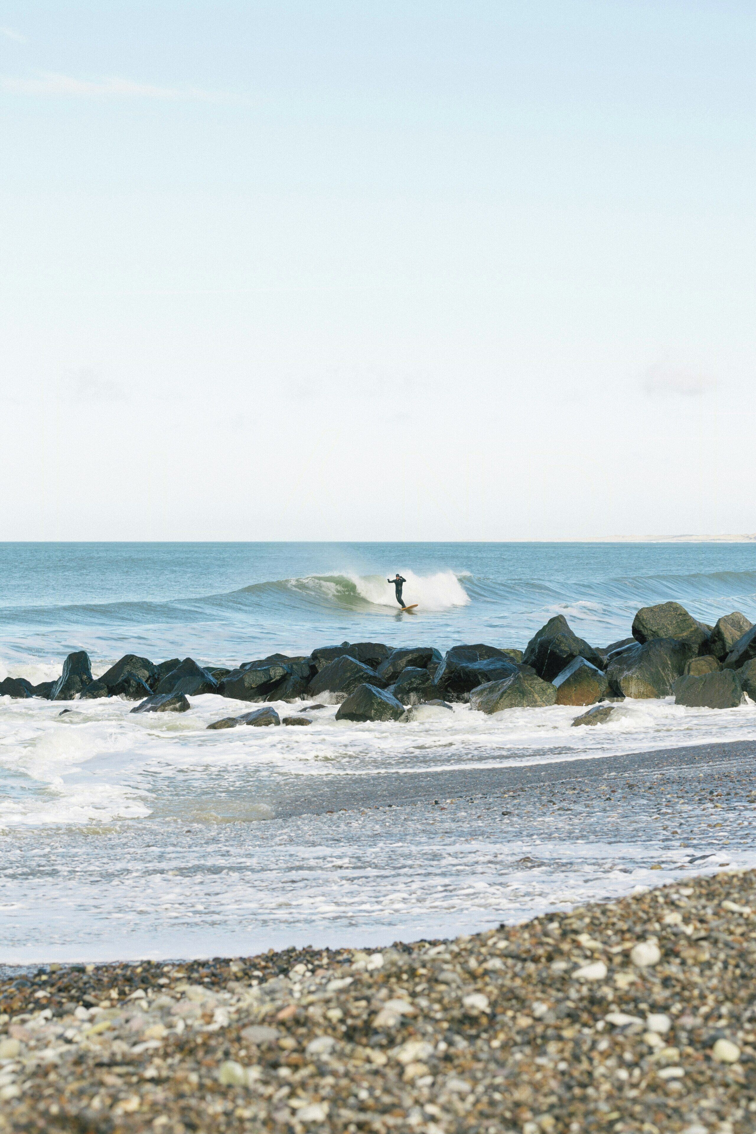 Una playa con rocas grandes, una persona practicando surf y mar tranquilo.