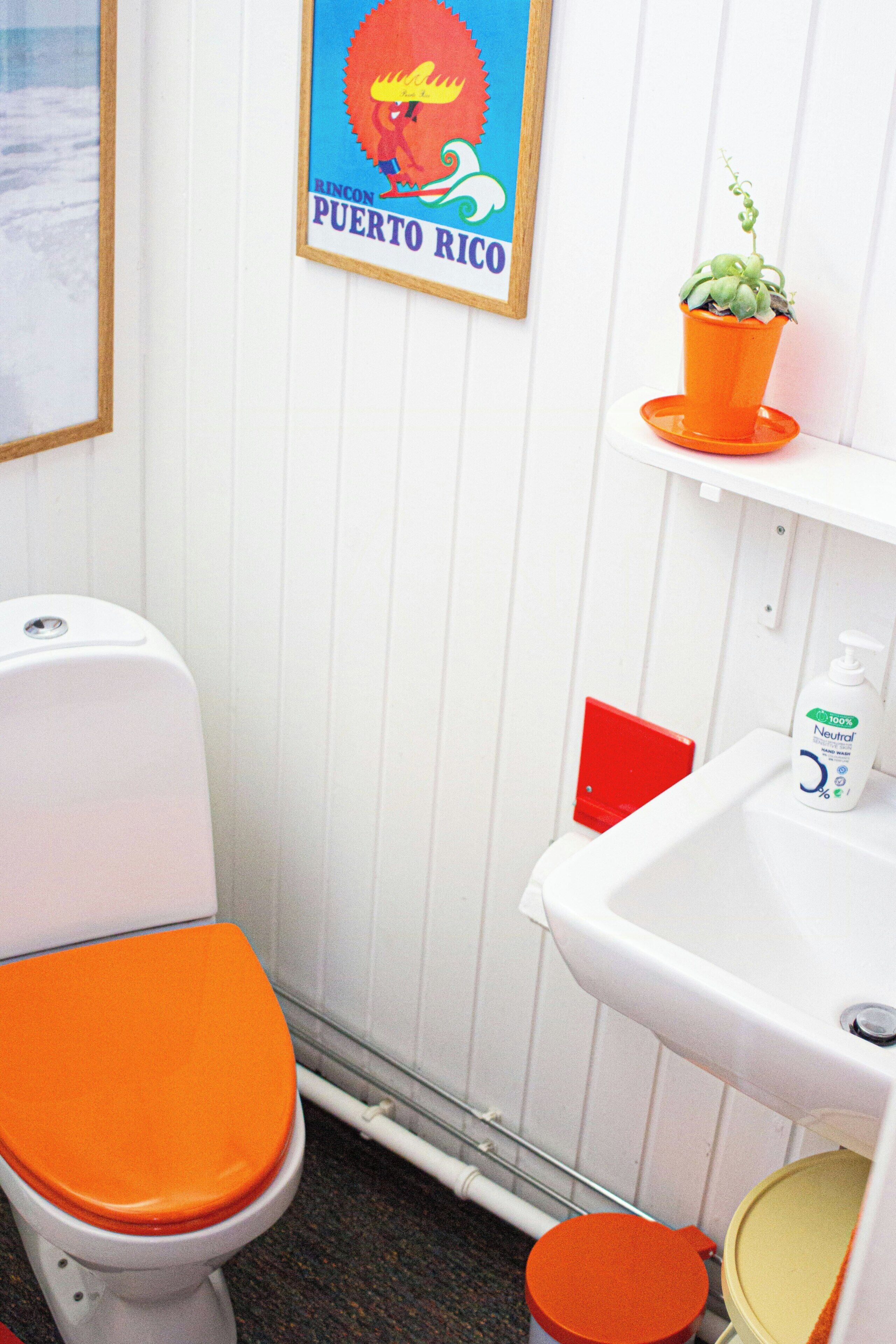 A bathroom with a white toilet, an orange seat, a sink with a bottle of hand soap, and a framed picture of Puerto Rico.
