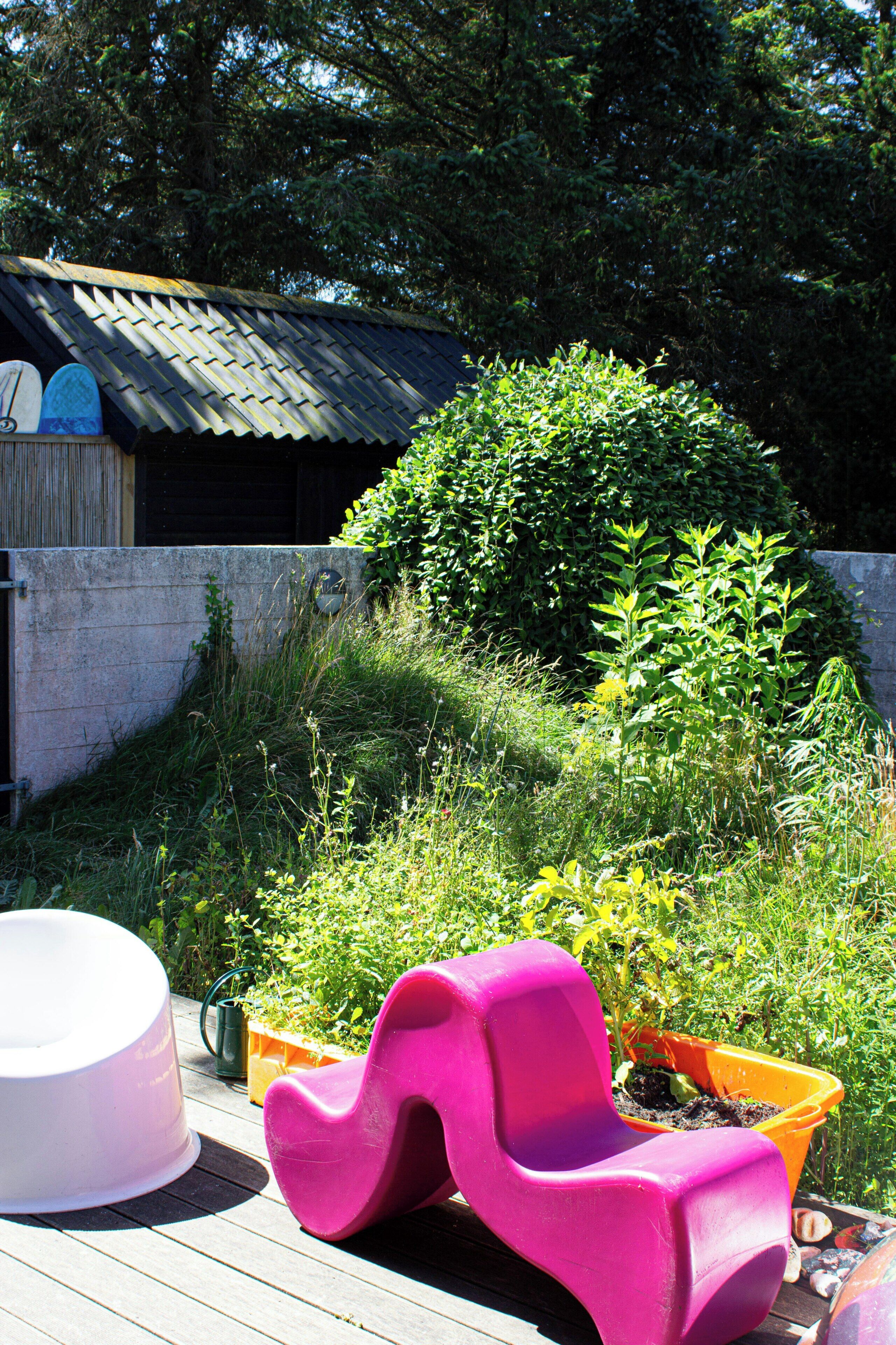 En træterrasse med en pink stol, en hvid plantekasse, og et lille haveområde med forskellige planter samt en metal konstruktion i baggrunden.