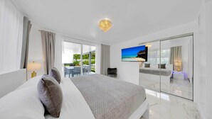 A modern hotel room with a large bed, a TV, a balcony with outdoor seating, and a view of the sea.