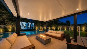 A modern outdoor living area with a pool, comfortable seating, and a large screen for entertainment.