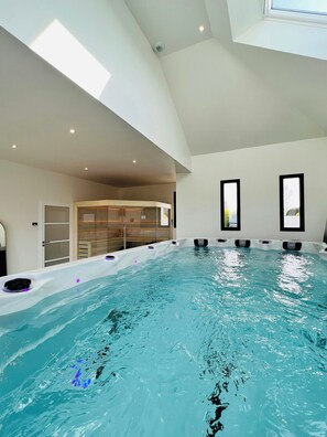 An indoor swimming pool with a high ceiling, built-in seating, and large windows. - Wellness villa with sea view in Brittany (Plouider)