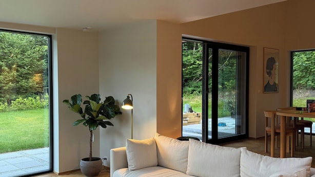 A modern living room with a white sofa, a glass coffee table, a potted plant, and large windows offering a view of a garden.