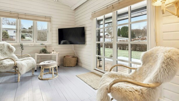 A room with white wooden walls, a flat-screen TV mounted on the wall, two fluffy chairs, a small table with a basket, and a large window with a view of a snowy landscape. - Comfortable summer house in Aabenraa (Aabenraa)