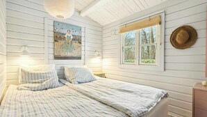 A bedroom with a bed, two bedside lamps, a painting, a window with a view, and a straw hat on the wall. - Comfortable summer house in Aabenraa (Aabenraa)