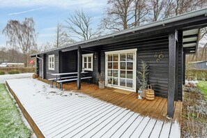 Eine schwarze Holzhütte mit einer Holzdiele, weißen Fenstern und einer kleinen Sitzgelegenheit im Freien.