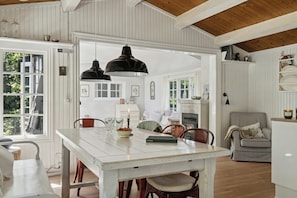 A dining area with a white table, chairs, and pendant lights. There is a view of a kitchen and a living area with a striped sofa. - Beach and forest close to Tisvilde house (Tisvildeleje)