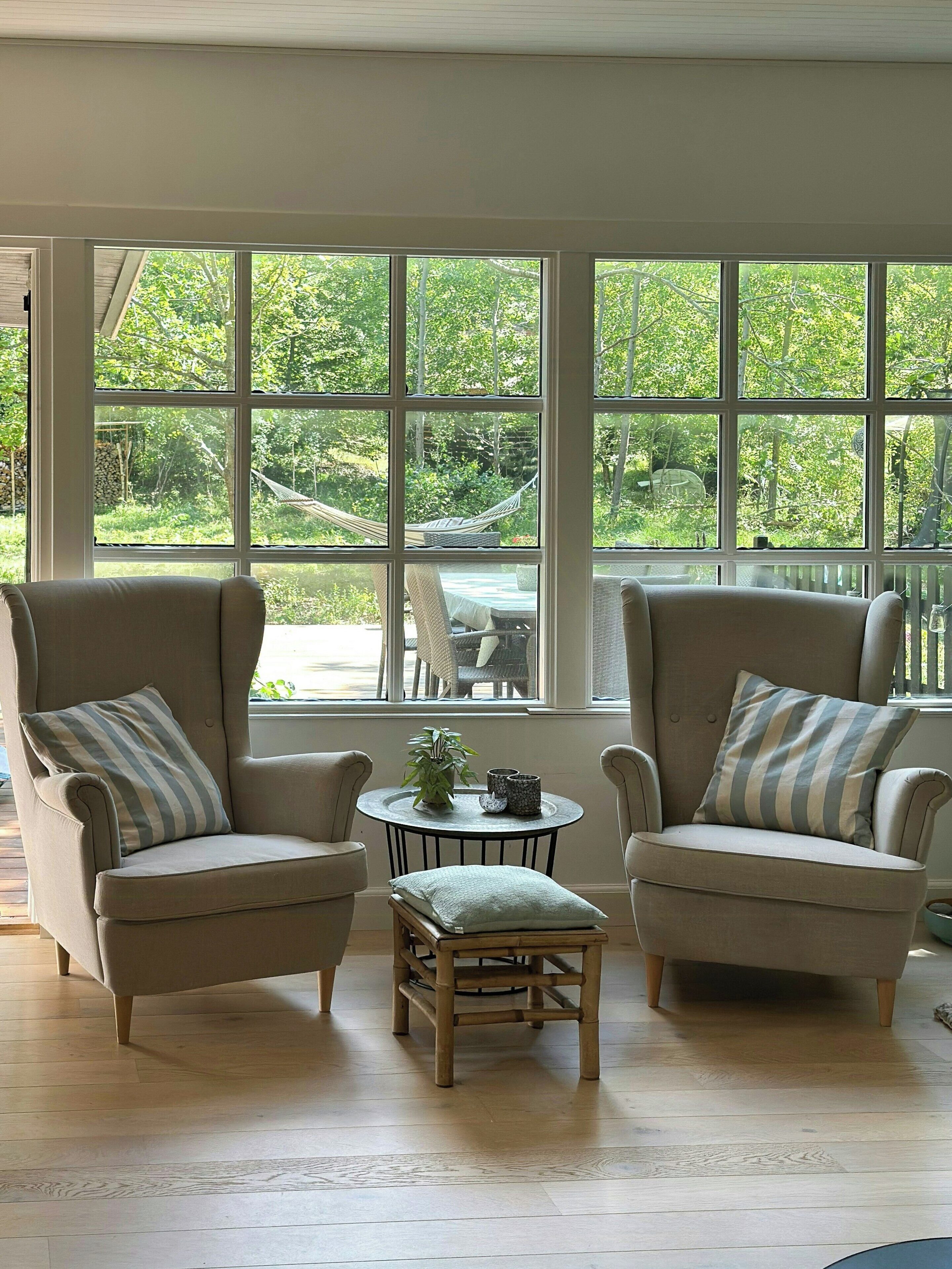 A room with two armchairs, a small table, and a view of a garden through large windows.