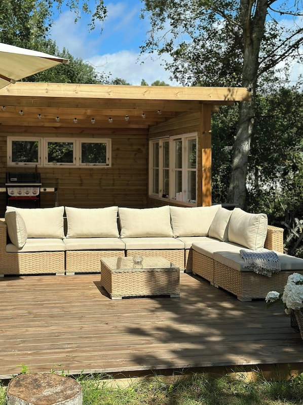Un terrazzo in legno con divano, poggiapiedi in vimini, barbecue e una pergola con luci a tendina.
