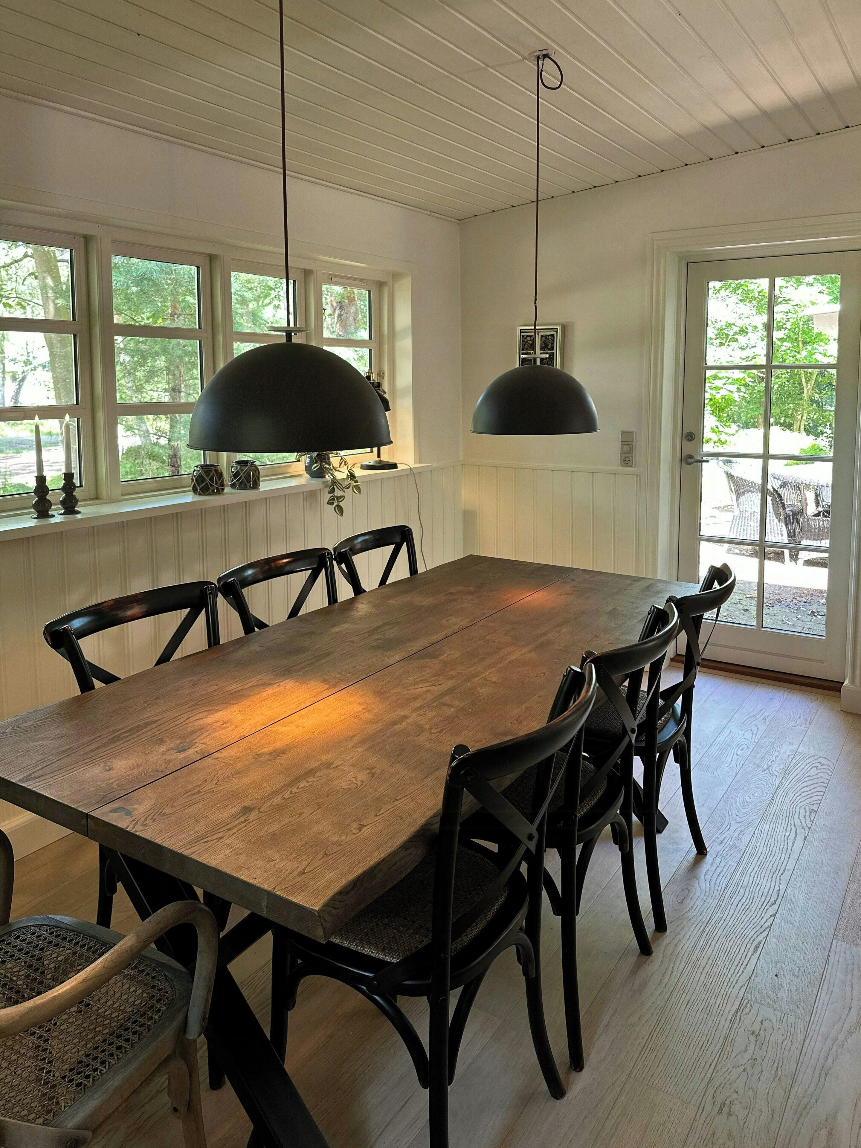 A dining room with a large wooden table, black chairs, pendant lights, and a view of greenery outside.