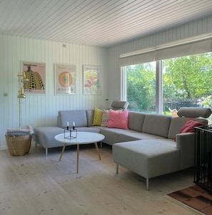 Ein modernes Wohnzimmer mit einem grauen Ecksofa, einem runden Couchtisch und einem großen Fenster mit Blick auf Bäume.