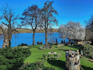 Un jardín junto al lago con una mesa y sillas de madera, un banco también de madera, y un tocón con un zapato.
