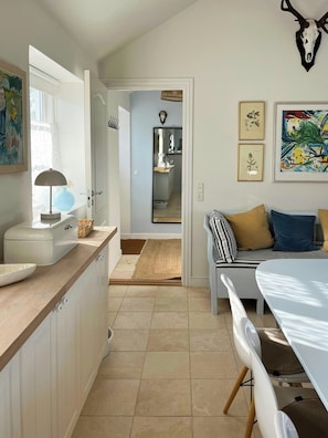 A modern kitchen with white cabinets, a dining table, and a kitchen island. There is a hallway leading to a living area with a sofa and a decorative skull on the wall. - House in peaceful surroundings 150 m to the water (Asperup)
