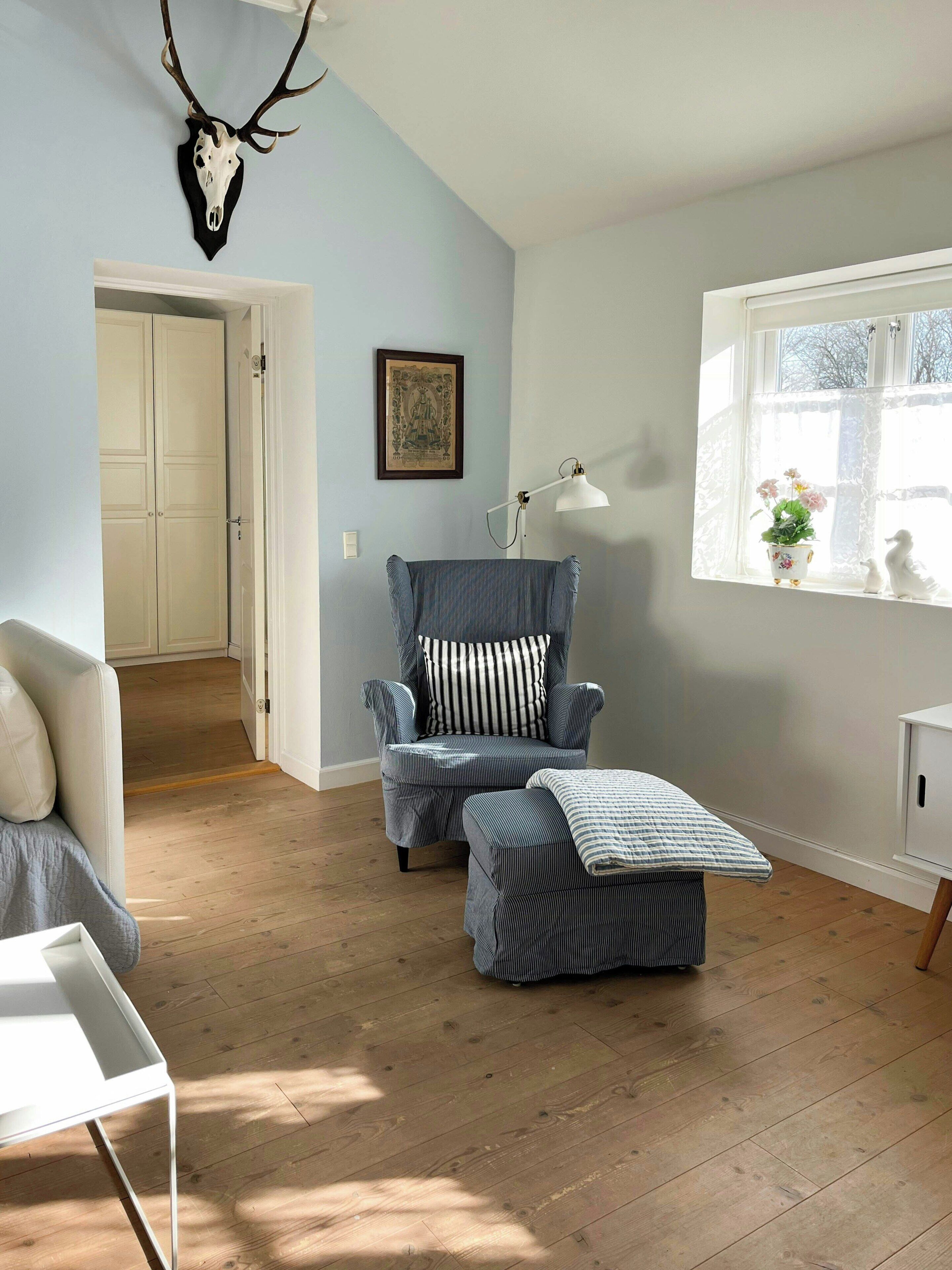 Une pièce avec un plancher en bois, un fauteuil à rayures, un canapé blanc, une fenêtre ornée de rideaux en dentelle et un crâne de cerf accroché au mur.