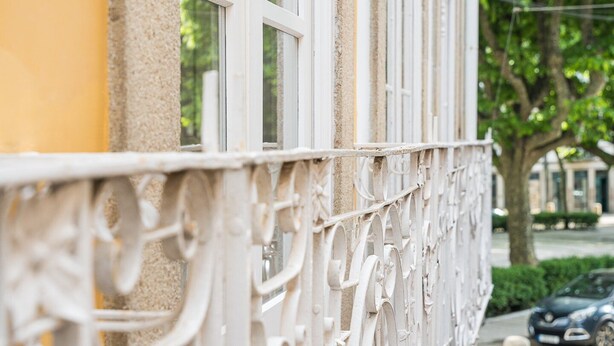 A white metal railing with intricate designs, a window, and a tree in the background.