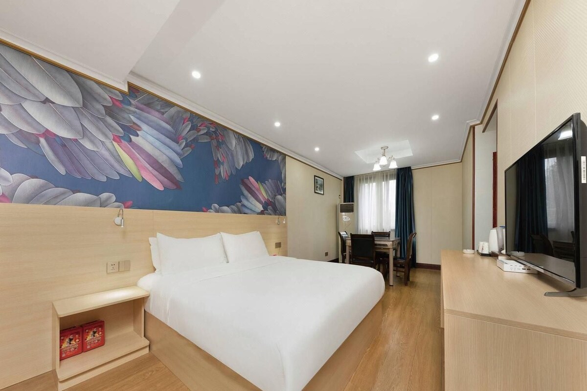 A hotel room with a large bed, a desk with a chair, a television, and a wall with a floral mural.