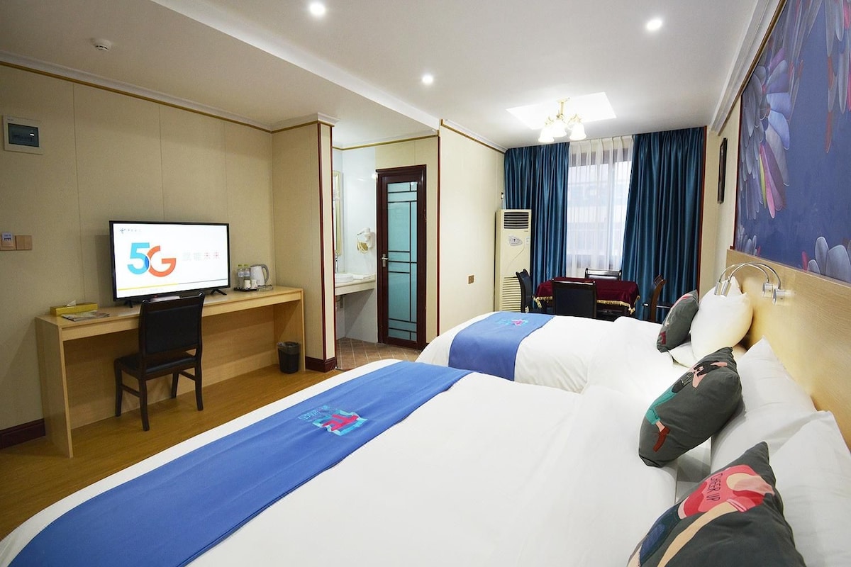 A hotel room with two beds, a desk with a computer, a television displaying '5G', and a bathroom door.
