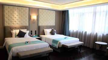 A hotel room with two beds, a bench, a bedside table with a lamp, a small round table, and a large window with curtains.