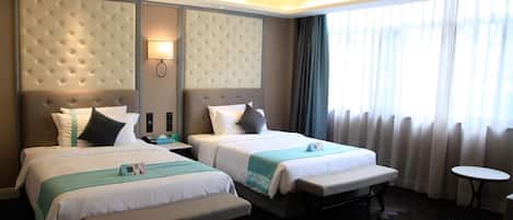 Habitación de hotel con dos camas, un banco, una mesita de noche con lámpara, una pequeña mesa redonda y un amplio ventanal con cortinas.