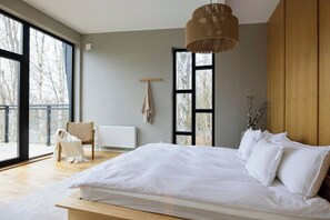 A bedroom with a large bed, a chair, a radiator, and a view of trees through the windows. - Modern forest villa with sea views and sauna (Trollhättan)