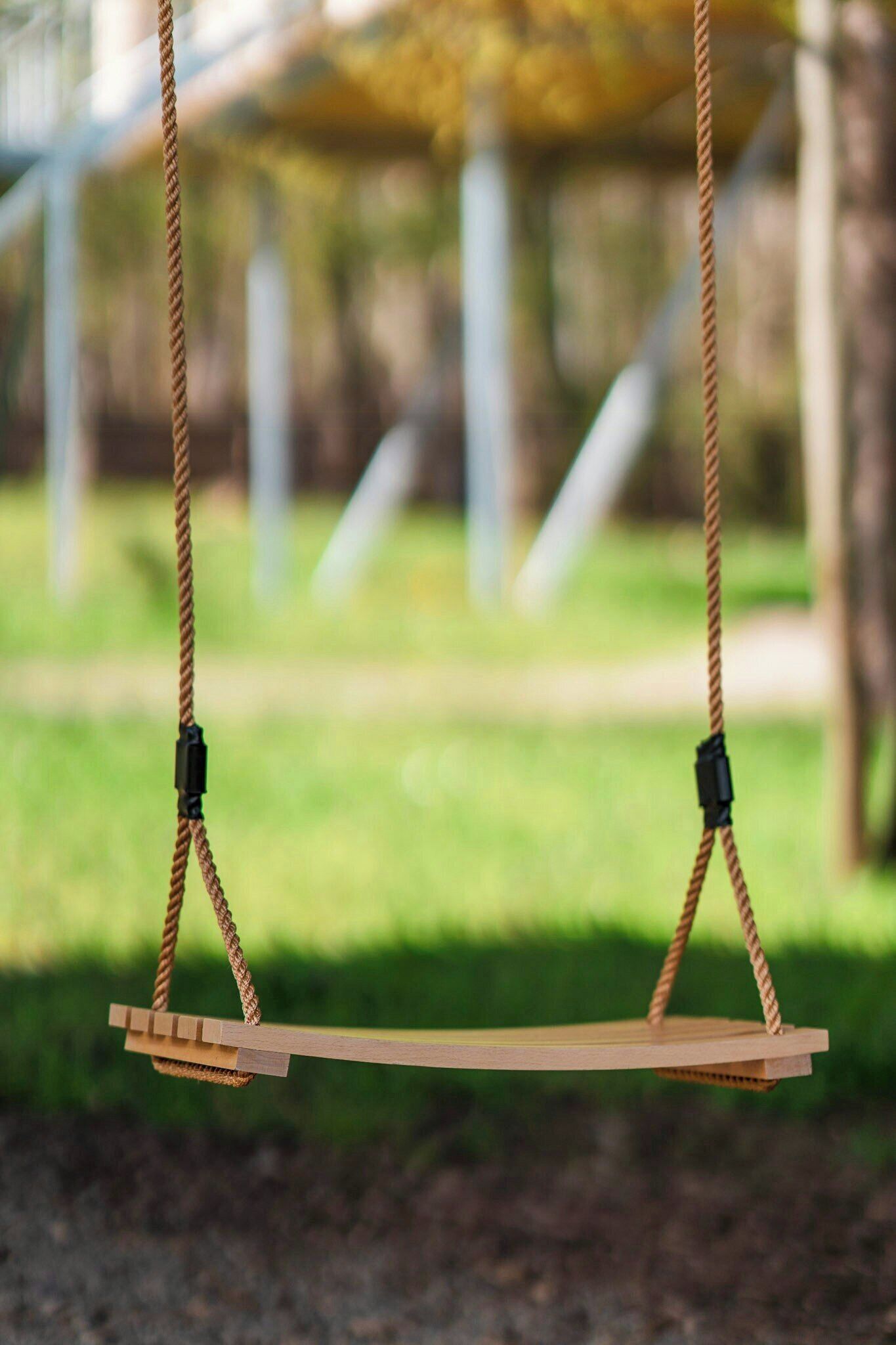 A wooden swing set with ropes and black clips, set against a blurred background of greenery and a structure.