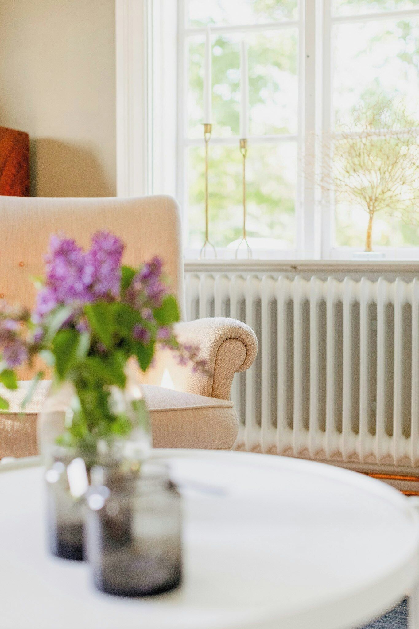 Ett rum med ett element, ett fönster med utsikt över grönområden, en beige fåtölj och ett vitt bord med en vas med blommor.