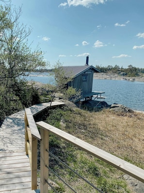 A wooden dock leading to a small cabin by a lake, surrounded by trees and rocky terrain. - Island house on private island with sea view (Östhammar)