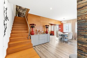 Ein modernes Interieur mit einer Holztreppe, einem Essbereich mit Tisch und Stühlen sowie einer Küchenzeile mit Schränken und einer Vase mit Blumen.