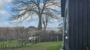 A grassy yard with a metal gate, a large tree, and a building with a dark exterior. - Cozy cottage with fjord view in Hjarbæk (Løgstrup)