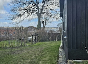 A grassy yard with a metal gate, a large tree, and a building with a dark exterior. - Cozy cottage with fjord view in Hjarbæk (Løgstrup)