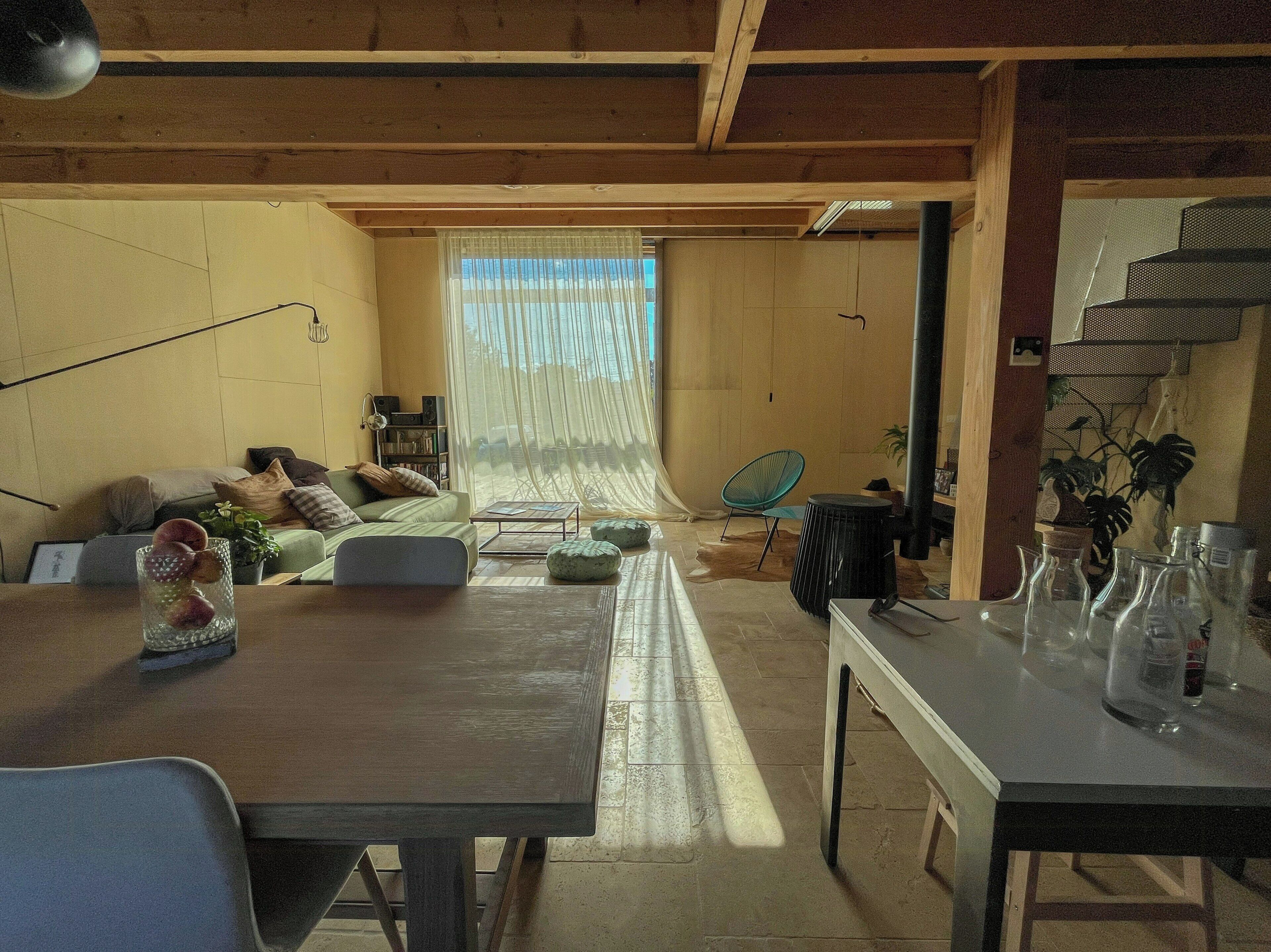 Een woonkamer met een bank, salontafel en een eettafel met stoelen. Er is een raam met dunne gordijnen en uitzicht op de omgeving.