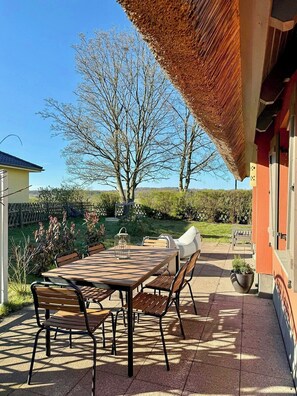 En terrasse med et bord og stole, et stråtækt tag og udsigt til træer og et hegn.