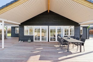 Ein modernes Gebäude mit dunkler Fassade, großen Glasüren und einer Holzterrasse mit Gartenmöbeln.