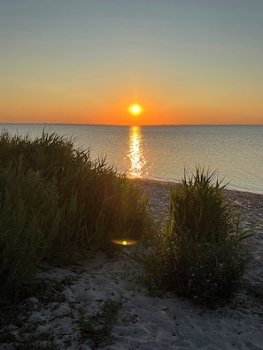 Ein Strand mit Sand, Gras und einem Sonnenuntergang über dem Wasser.