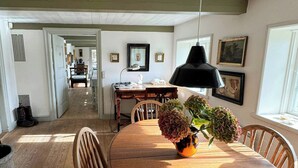 A dining area with a wooden table, chairs, a vase with flowers, a pendant light, and a view of a kitchen and hallway. - Protected farm from 1798 by Blåvand beach (Blåvand)