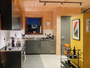 A modern kitchen with wooden cabinets, a built-in oven, a sink, and a window with a view of fireworks. - Wooden home among apple trees with ocean view (Kivik)
