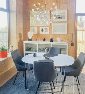 A modern dining area with a round table, four chairs, a white cabinet with glass doors, and a wooden wall with framed pictures. - Wooden home among apple trees with ocean view (Kivik)