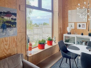 A modern room with a large window, wooden panel walls, a dining area with chairs, and a small shelf with decorative items. - Wooden home among apple trees with ocean view (Kivik)