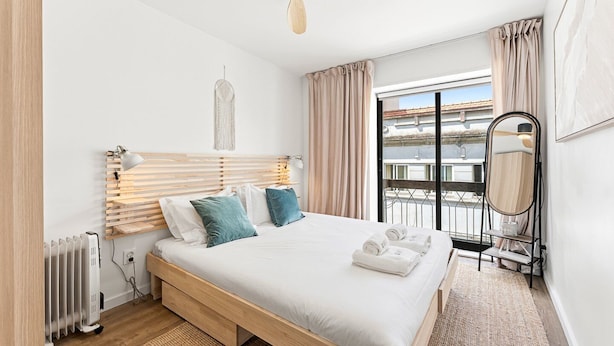 Una habitación de hotel moderna con una cama de madera, un ventanal con vistas, un espejo de pie y un radiador.