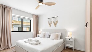 Una habitación de hotel moderna con una cama grande, una mesita de noche con lámpara, un ventilador de techo y vistas a la ciudad desde la ventana.
