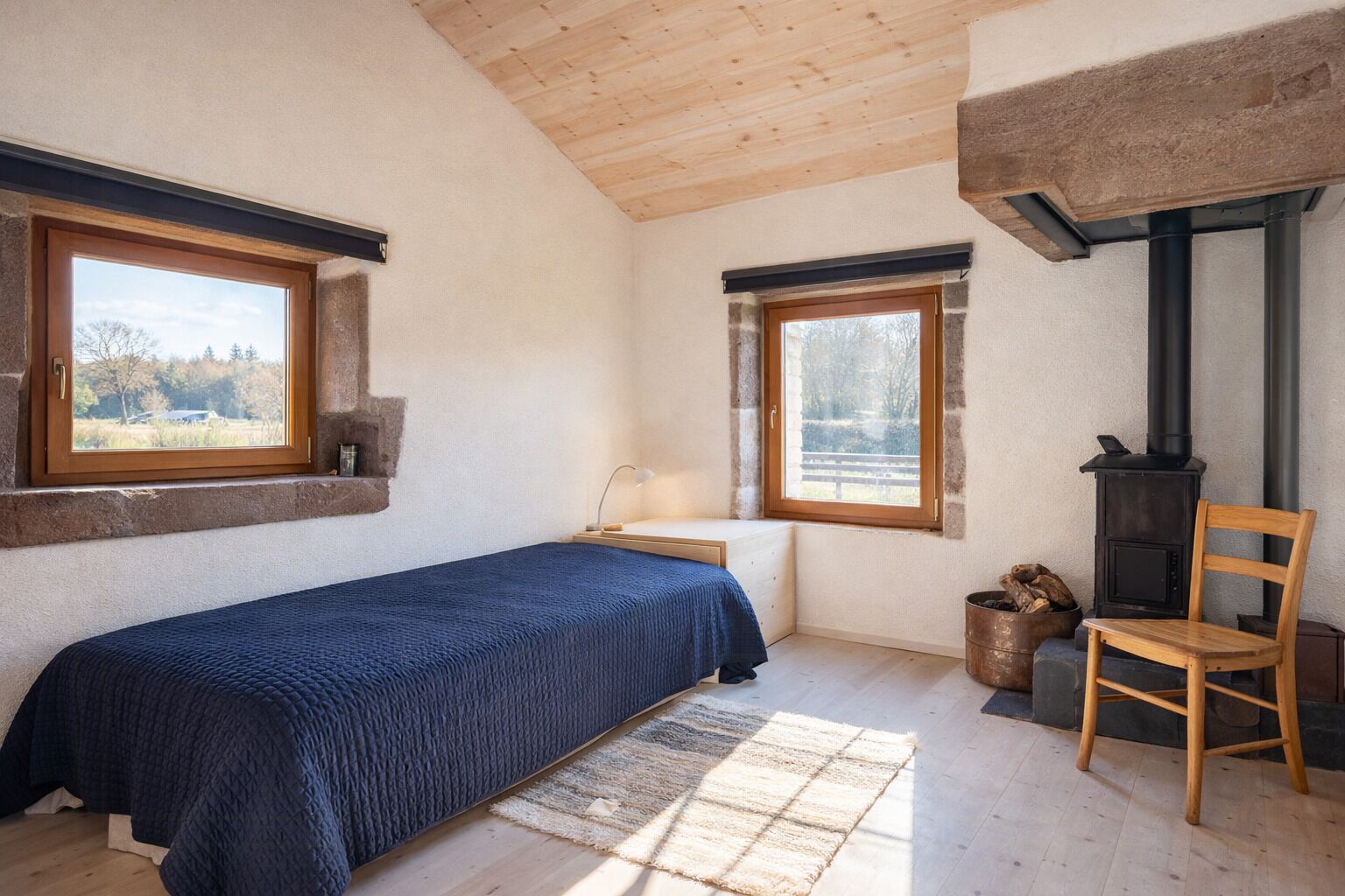 Un dormitorio con una cama, dos ventanas, una silla de madera, una chimenea y vistas al exterior.