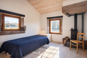 Ein Schlafzimmer mit einem Bett, zwei Fenstern, einem Holzstuhl, einem Kamin und Blick ins Freie.