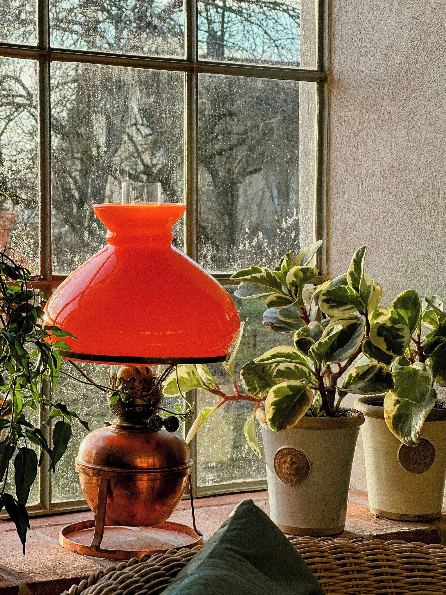 Et værelse med et stort vindue, en kobber gryde på et stativ, en rød lampe og flere plantede planter.
