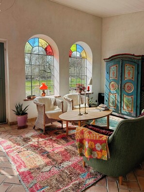 Ein Zimmer mit großem Fenster, einem grünen Sessel, einem runden Tisch und einem Schrank mit floralem Dekor.