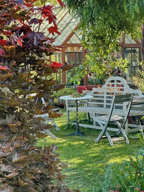 Ein Garten mit einer hölzernen Bank und einem Tisch, umgeben von viel Grün und einem Gewächshaus.