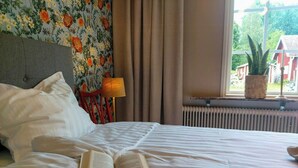 Un lit avec un livre, une lampe et une fenêtre donnant sur une maison et de la verdure.