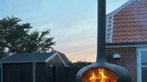 En brændefyret pizzaovn med skorsten, et stabel med brænde og et murstenshus i baggrunden.