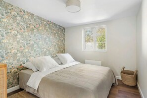Ein Schlafzimmer mit blumigem Wandpapier, einem Bett mit weißer Bettwäsche, einem Fenster mit Blick und einem Korbwäschekorb.
