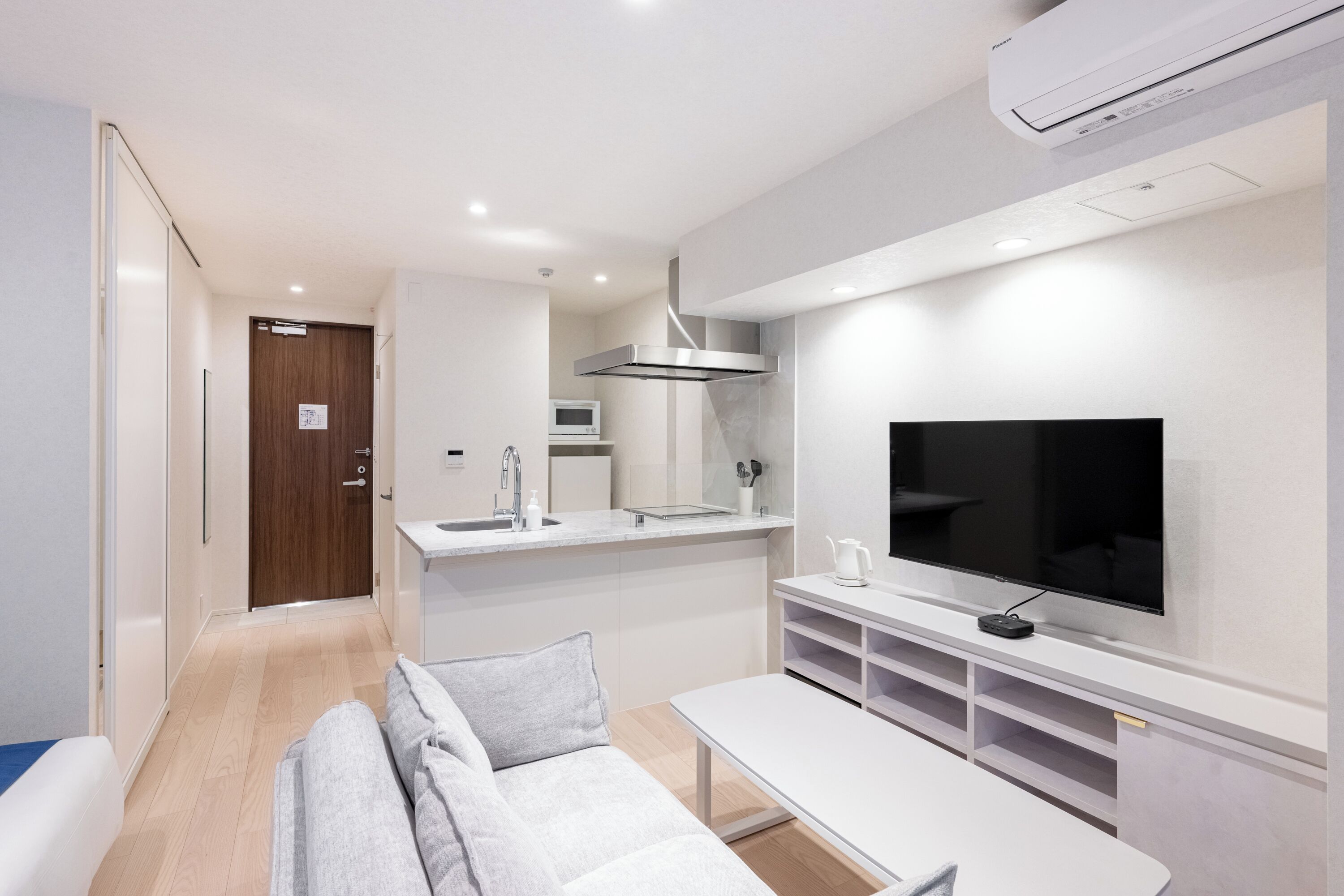 A modern hotel room with a sofa, a dining table, a TV, a kitchen area, and an air conditioning unit.